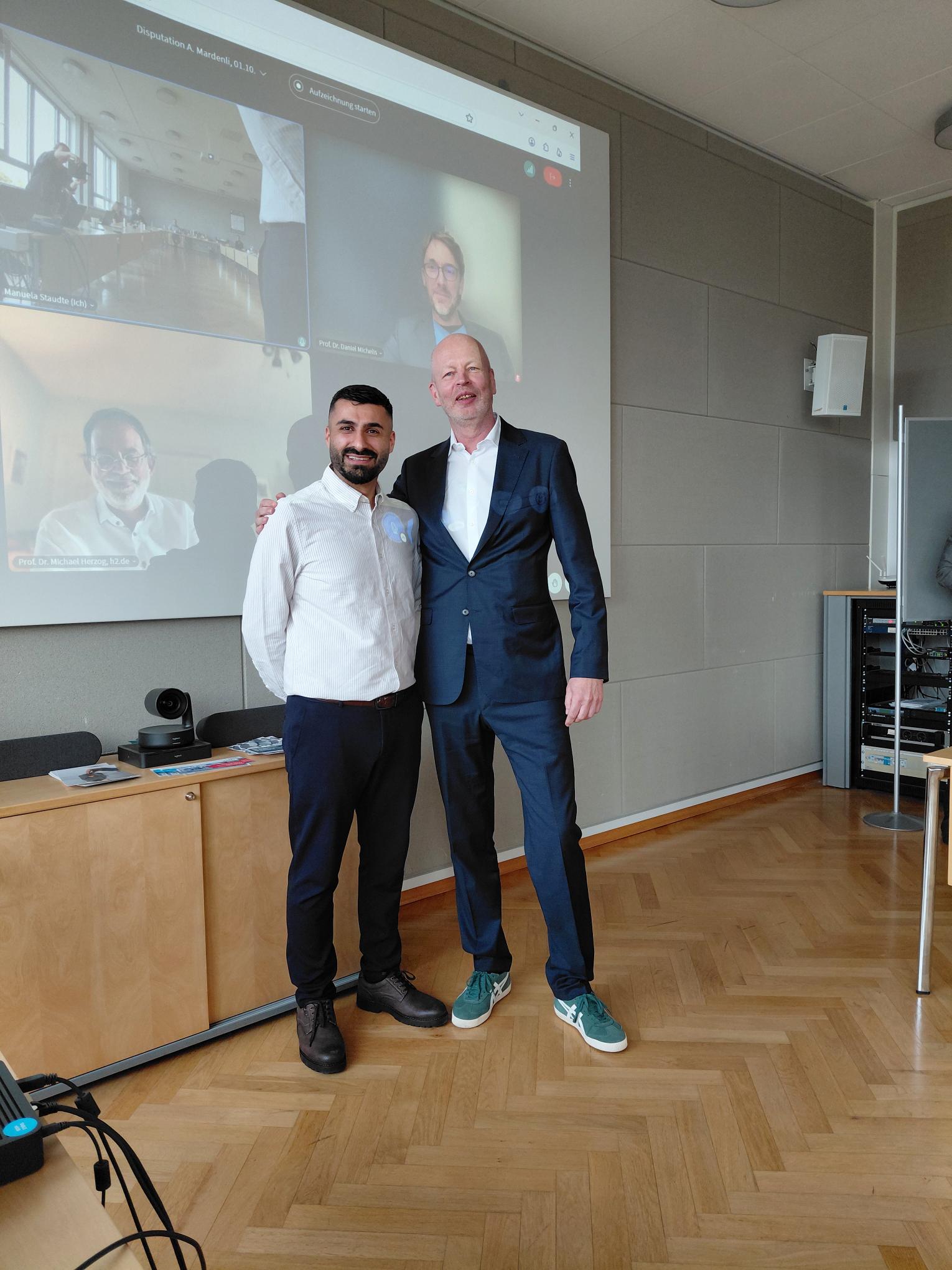 Abdulaziz Mardenli schließt als erster Promovierender seine Promotion am Promotionszentrum für Sozial-, Gesundheits- und Wirtschaftswissenschaften auf dem Fachgebiet der Wirtschaftswissenschaften ab 2 Personen stehen nebeneinander