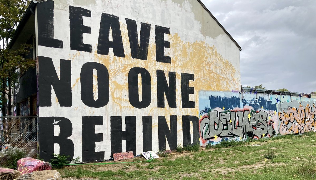 Eine Hausfassade, auf die der Schriftzug "Leave No One Behind" aufgemalt wurde. Rechts schließt sich eine Mauer mit bunten Graffitis an. 