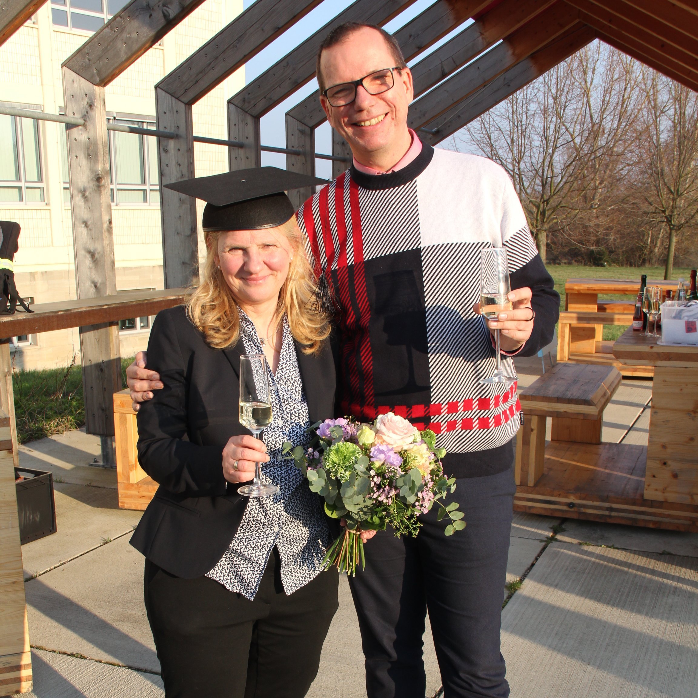 Disputation_Anja_Jonas_19.3.26 Auf dem Bild sind zwei Personen zu sehen. Links eine Frau mit Blumenstrauß und Doktorhut, rechts ein Mann mit einem Sektglas in der Hand