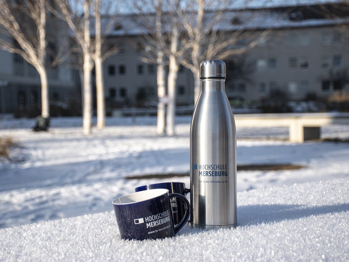 Hochschulshop: Tassse und Isolierflasche Eine blaue Tasse neben einer silbernen Isolierflasche mit Hochschullogo im Schnee