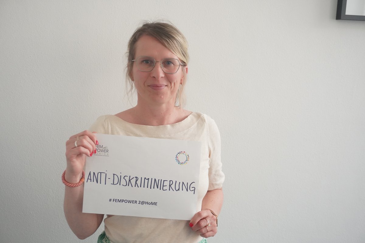 Person steht vor einer weißen Wand mit einem Schild Eine weiblich gelesene Person steht vor einer weißen Wand und trägt ein Schild mit dem Aufdruck Anti-Diskriminierung