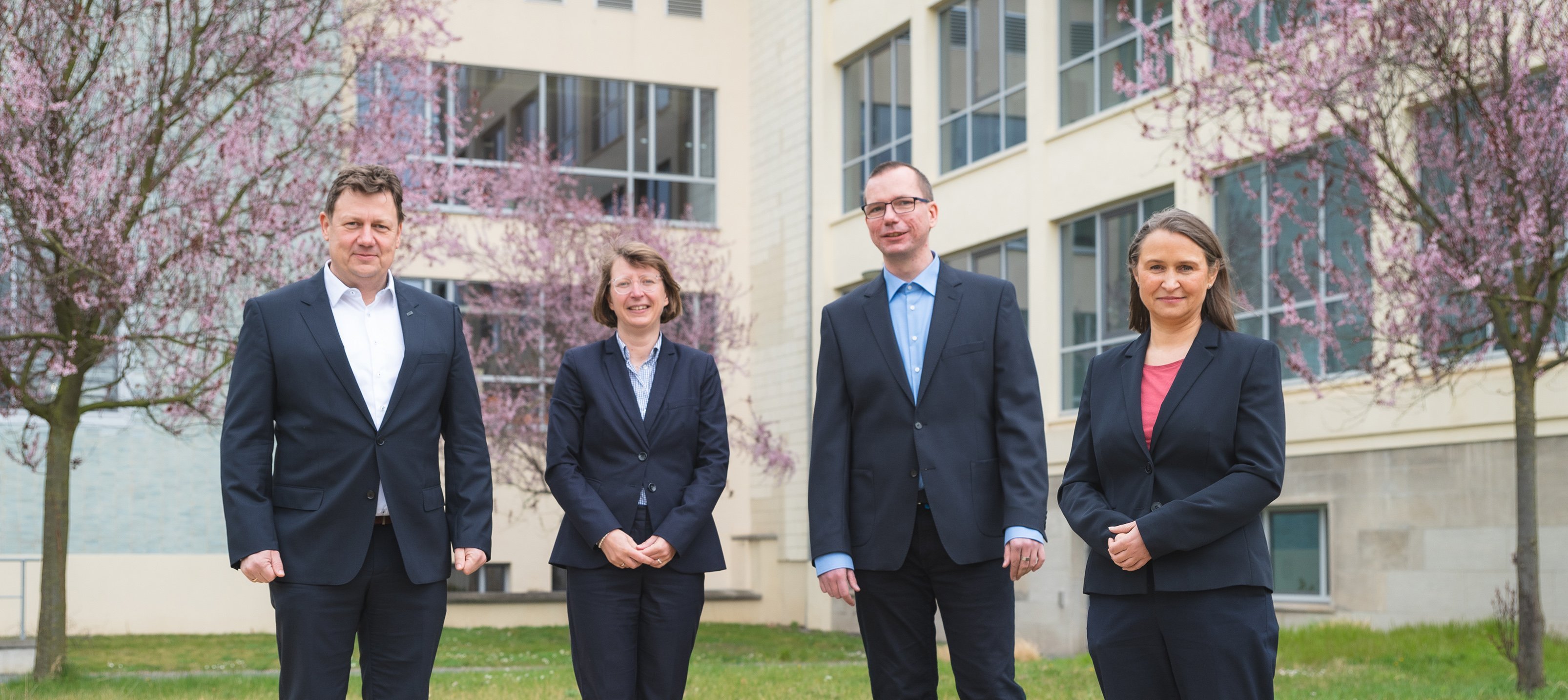 Gruppenbild Rektorat Vier Personen im Freien im Anzug im Hintergrund ein Gebäude mit großen Fenstern