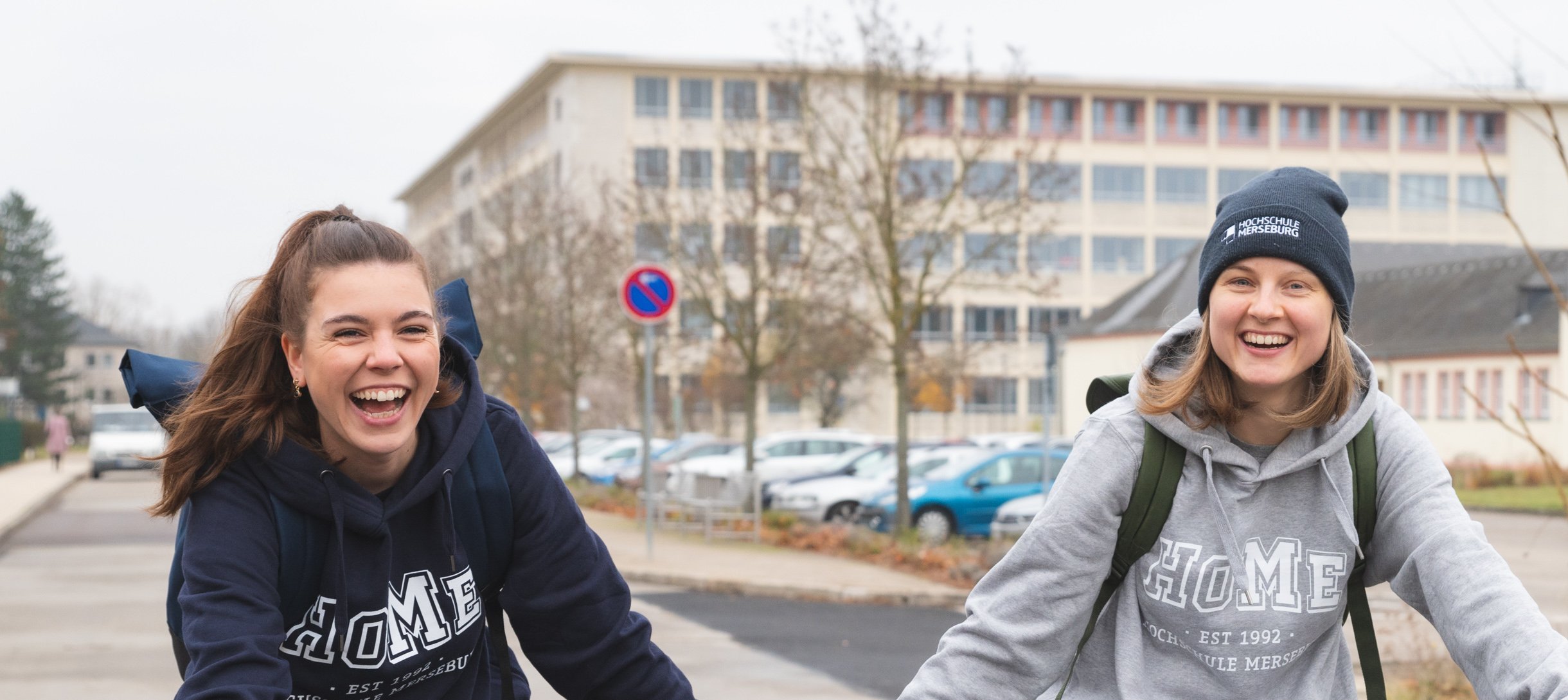 Hochschulshop Fahrrad 2 Personen lachend mit blauen und grauen Hochschulpullover an auf Fahrrädern im Vordergrund, im Hintergrund ein großes Gebäude mit Fenstern, Jahreszeit ist Herbst
